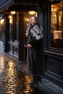 A woman stands on a wet cobblestone street outside a dark storefront at night, wearing a long dark fur coat with a contrasting light gray fur collar and black boots.