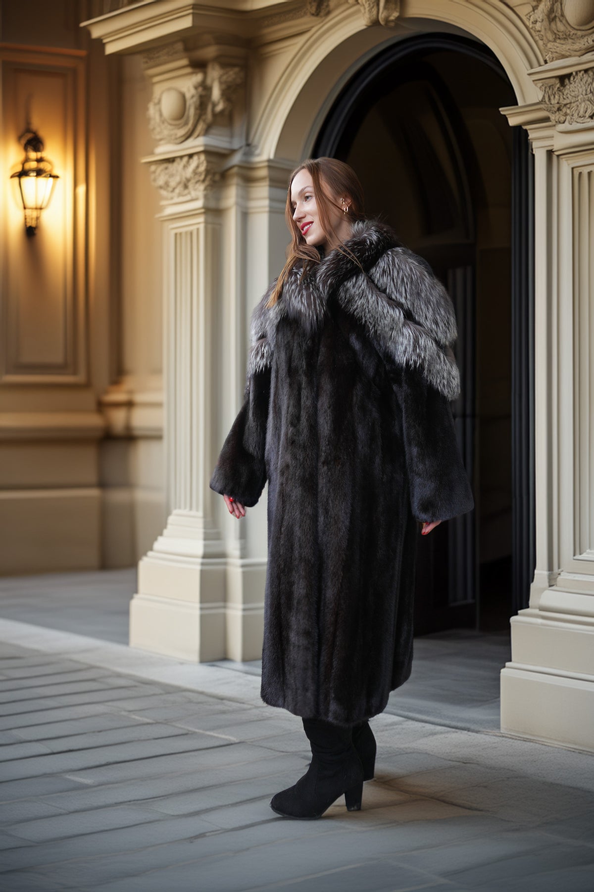 A woman wearing a long black fur coat with a gray and white fur collar, standing in front of an ornate stone doorway.