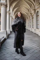 A woman wearing a long black fur coat with a gray and white fur collar, standing in a grand arched corridor with stone columns and tiled floors.