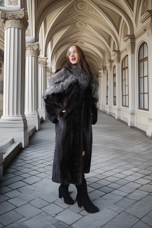 A woman wearing a long black fur coat with a gray and white fur collar, standing in a grand arched corridor with stone columns and tiled floors.