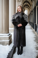 A woman wearing a long dark gray fur coat with a large silver-gray and black fur collar, standing in a snowy outdoor corridor with classical columns.
