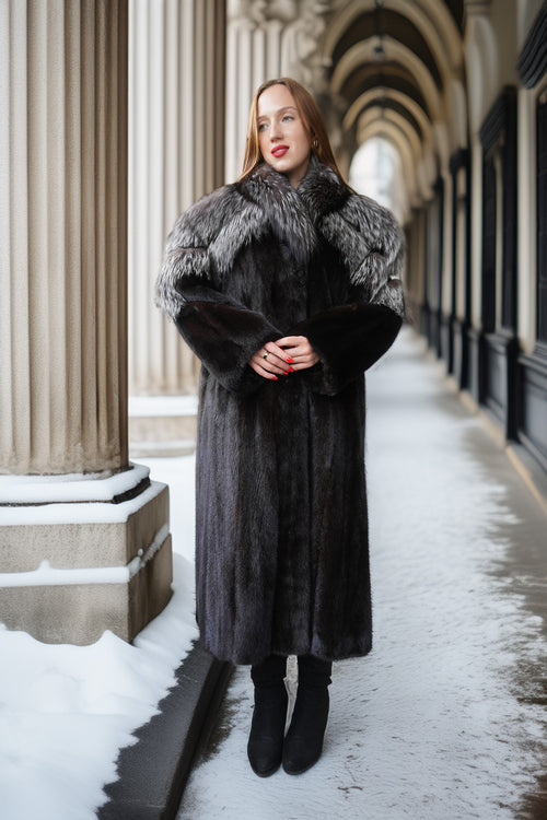 A woman wearing a long dark gray fur coat with a large silver-gray and black fur collar, standing in a snowy outdoor corridor with classical columns.