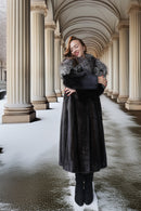 A woman wearing a long dark fur coat with a gray fur collar stands in a snowy colonnade between large beige columns.