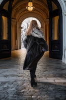 A woman viewed from behind wearing a long dark faux-fur coat with a light gray fur collar, walking through a grand stone corridor with arched doorways and a hanging lamp.
