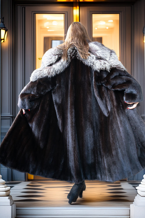 A woman viewed from behind wearing a voluminous dark fur coat with a wide light-gray fur collar, standing on a checkered porch in front of a grand doorway.