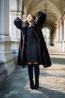 A woman posing in a long black fur coat with a dark dress and over-the-knee boots, standing in a sunlit classical stone corridor.