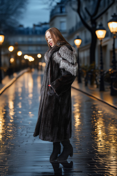 A woman wearing a long dark fur coat with a light gray fur collar stands on a wet cobblestone street at dusk, with glowing street lamps and blurred buildings in the background.