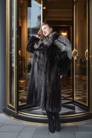 A woman wearing a long black fur coat with a large gray fur collar stands in front of a luxury revolving door entrance.