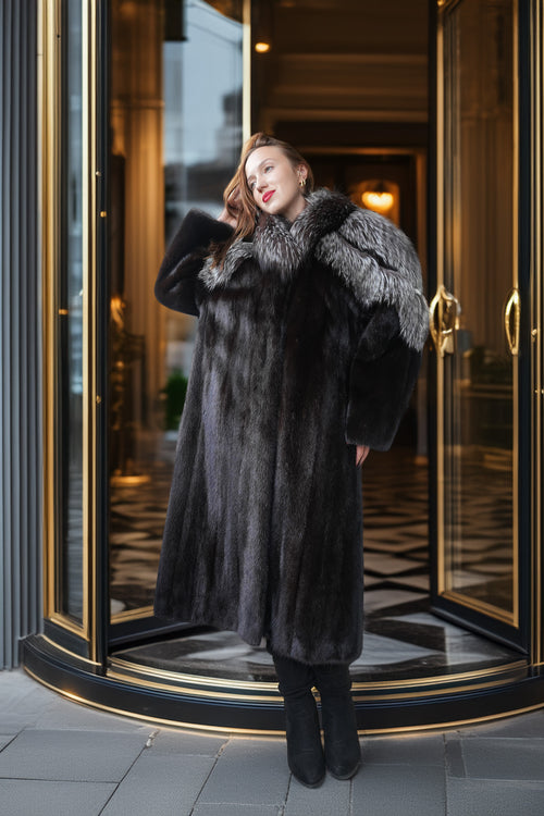 A woman wearing a long black fur coat with a large gray fur collar stands in front of a luxury revolving door entrance.
