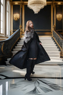 A woman posing on a grand marble staircase in a luxurious interior, wearing a long black coat with a gray fur collar, a black dress, and black knee-high boots.