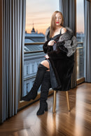 A woman sits on a chair in a luxurious room by a large window, wearing a black faux fur coat with gray fur trim, black thigh-high boots, and a black dress. The background shows a cityscape at sunset with gray curtains framing the window.
