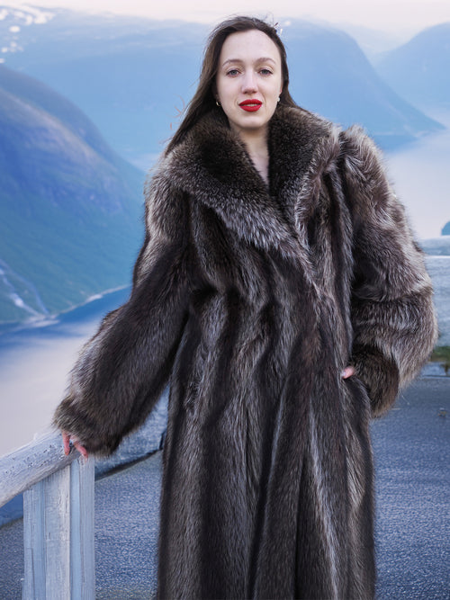 Front view of woman in silver-tipped raccoon fur coat, standing on balcony with fjord view, showcasing luxury outerwear.