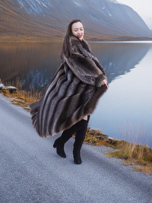 Model twirling in full-length raccoon fur coat beside lake, showcasing movement and plush texture of luxury winter outerwear.