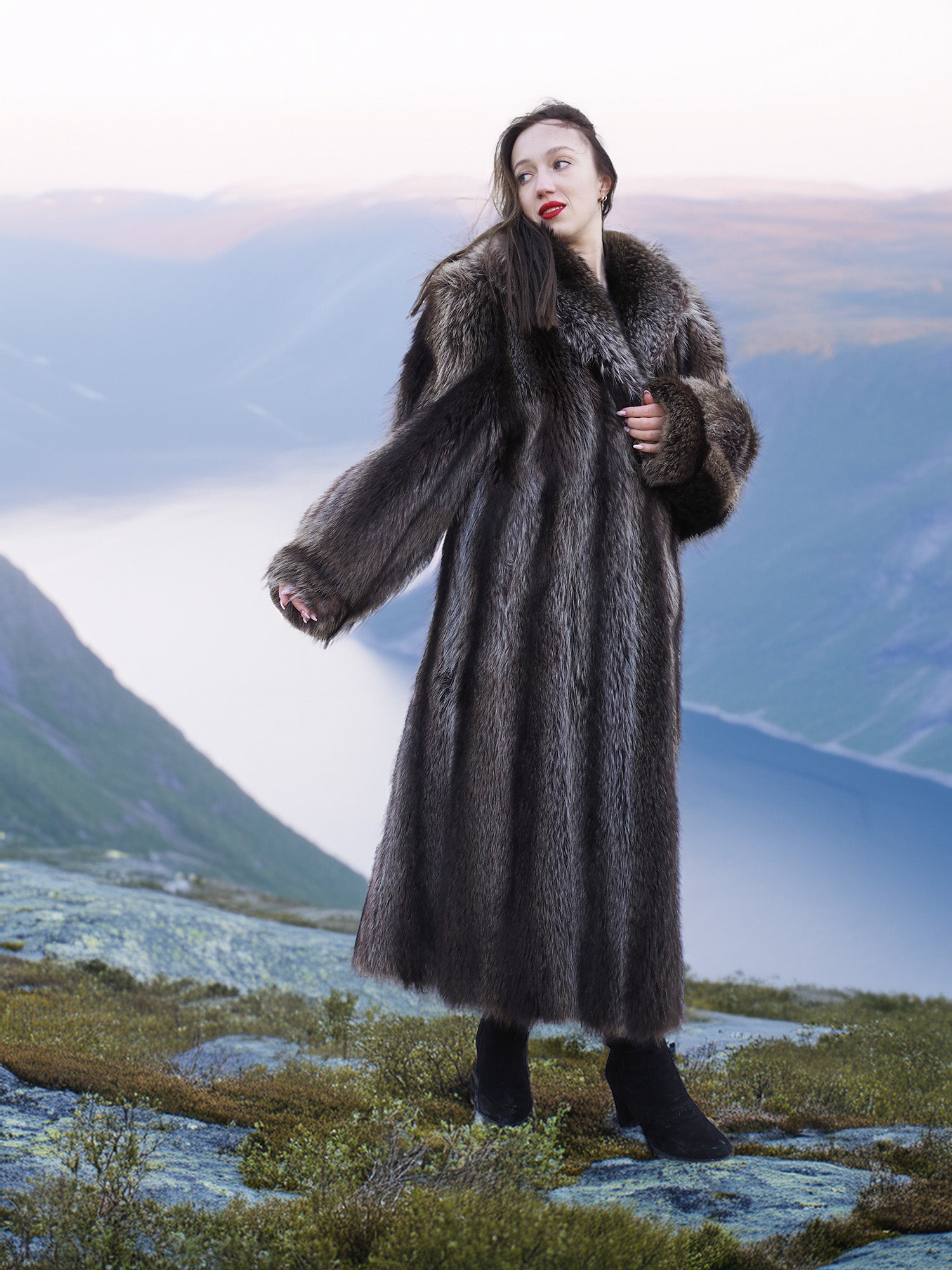 Model in raccoon fur coat standing on rocky cliff with fjord view, showcasing natural fur elegance and winter outerwear.