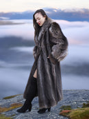 Model in full-length raccoon fur coat walking on rocky terrain above clouds, highlighting natural fur elegance and warmth.