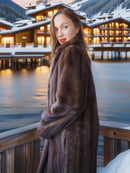 Woman in a fur coat standing in front of a snowy mountain resort at dusk.