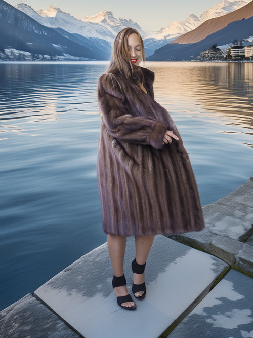 Woman in a fur coat standing by a lake with mountains in the background