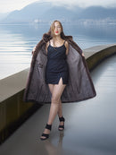 Woman in a long coat standing on a dock with water and mountains in the background