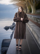Woman in a fur coat standing by a lake with mountains in the background