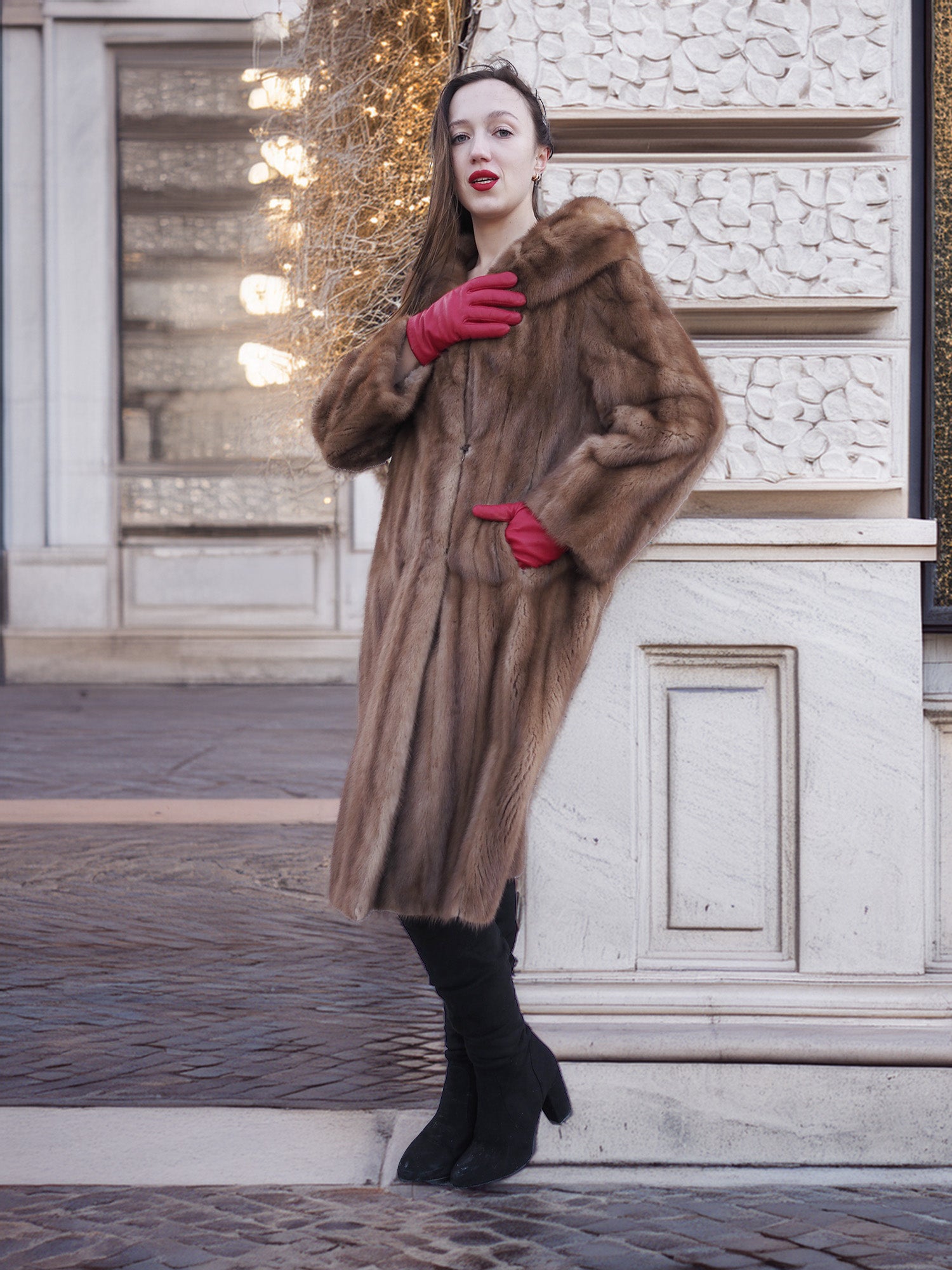 Model leaning on wall in vintage pastel Canadian brown mink fur coat, styled with red gloves and boots for timeless cold-weather elegance.