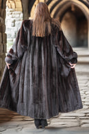 Person wearing a long dark fur coat walking away from the camera in an outdoor setting with stone architecture.