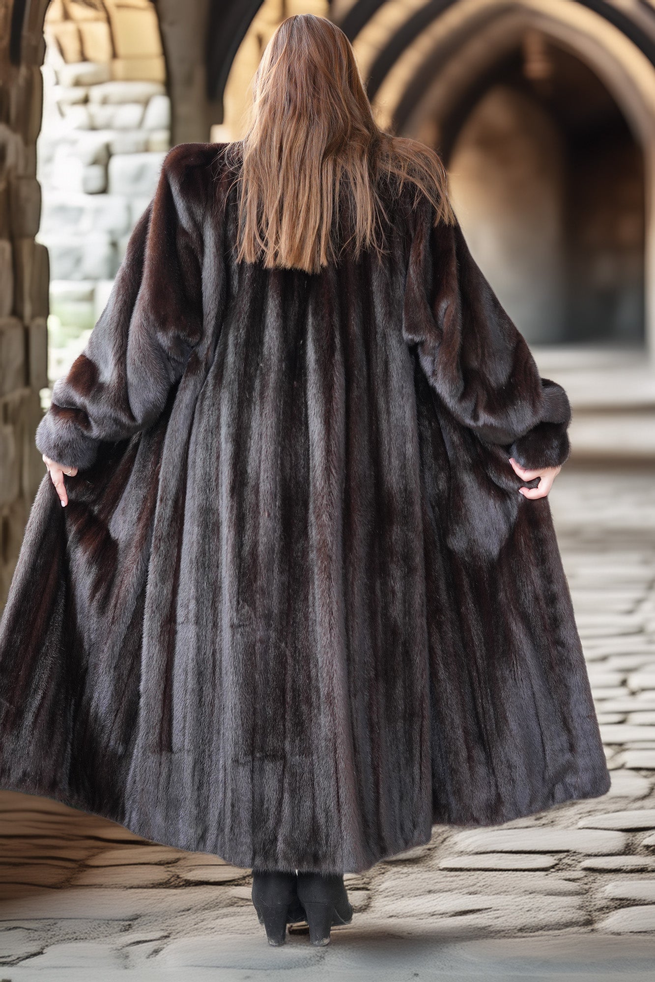 Person wearing a long dark fur coat walking away from the camera in an outdoor setting with stone architecture.