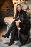 Woman in a dark fur coat and black boots sitting against a stone wall.