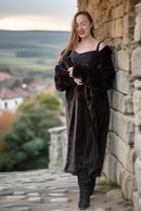 Woman in a long dark coat standing against a stone wall with a scenic background
