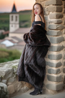 Woman in a fur coat standing against a stone wall with a scenic background