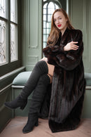 Woman wearing a dark fur coat sitting in a room with large windows.