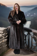 Woman in a fur coat standing on a balcony with a scenic view of a river and mountains.