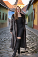 Woman in a long coat and black outfit standing on a cobblestone street.