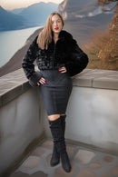 A woman posing on a stone balcony overlooking a scenic lake and mountains, wearing a black faux fur cropped jacket, a black bodycon pencil skirt with a thigh cutout, and black over-the-knee boots.