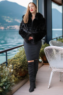 A woman stands on a balcony overlooking water and mountains, wearing a black faux-fur cropped jacket, a dark gray fitted midi skirt with a thigh cutout, and black over-the-knee boots.