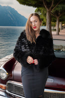 A woman stands outdoors beside a vintage maroon car, wearing a black patterned faux fur wrap or jacket over a dark gray skirt. Lake water, trees, and mountains are visible in the background.