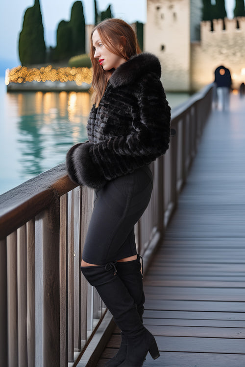 A woman stands on a wooden boardwalk by the water wearing a black faux fur cropped jacket, black leggings, and black over-the-knee boots, with a castle and water in the background.