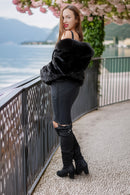 A woman stands on a lakeside walkway wearing a voluminous black faux-fur jacket, black ribbed pants, and black over-the-knee boots, with a metal railing and distant mountains in the background.