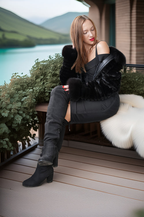 A woman sits on a wooden bench on a deck, wearing a black off-the-shoulder dress, a black faux-fur jacket, black over-the-knee boots, and holding a black shoulder bag. A scenic lake and mountains are visible in the background.