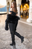 A woman in a black off-the-shoulder top, black pants, and black over-the-knee boots stands on a cobblestone walkway in an outdoor architectural setting, holding a black faux-fur wrap around her arms.