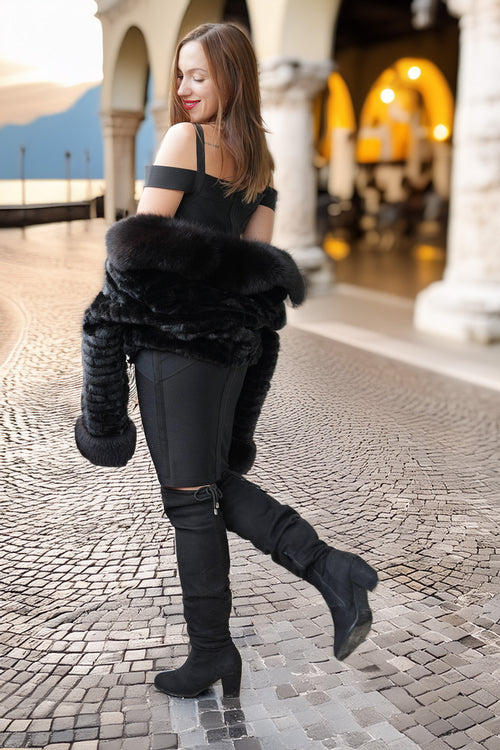 A woman in a black off-the-shoulder top, black pants, and black over-the-knee boots stands on a cobblestone walkway in an outdoor architectural setting, holding a black faux-fur wrap around her arms.