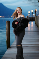 A woman stands on a wooden dock by the water at dusk, wearing a black off-the-shoulder top, dark gray fitted pants, black thigh-high boots, and holding a black faux-fur accessory. Mountains and lake are visible in the background.