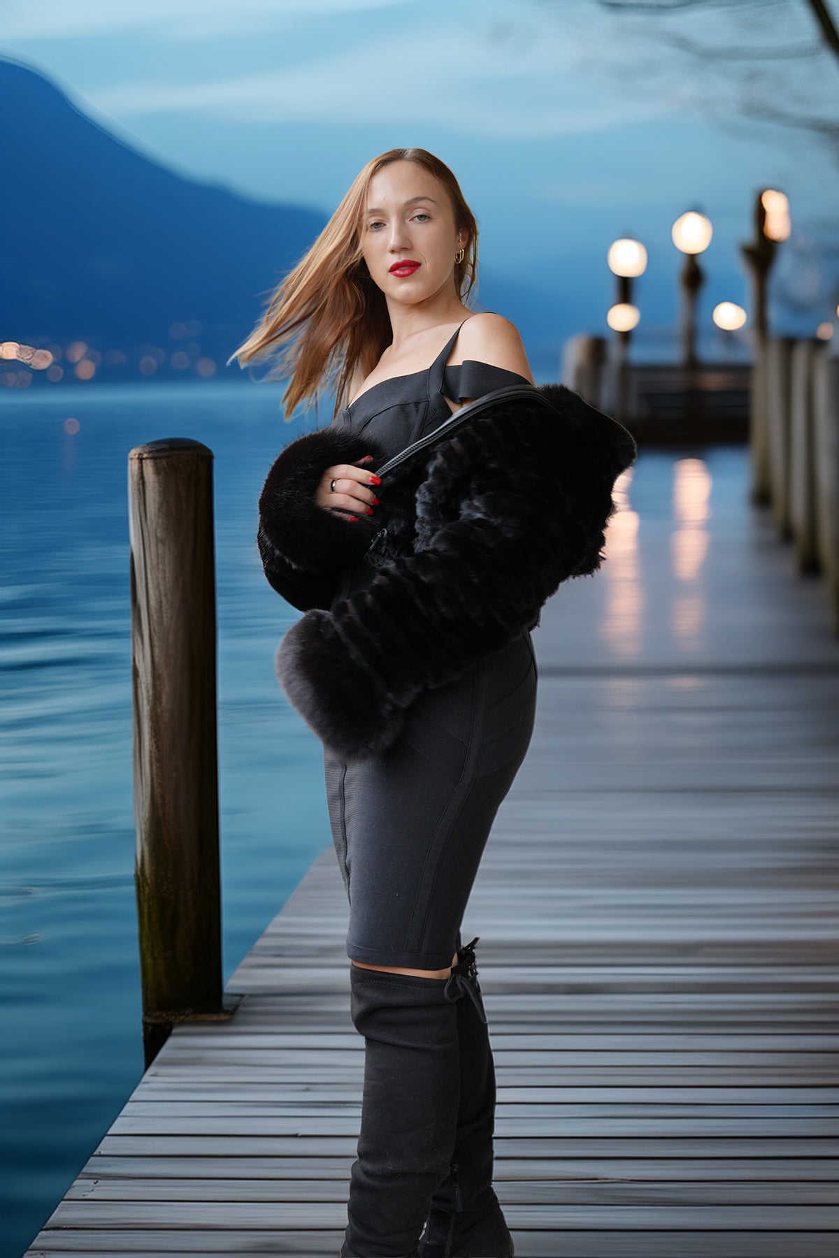 A woman stands on a wooden dock by the water at dusk, wearing a black off-the-shoulder top, dark gray fitted pants, black thigh-high boots, and holding a black faux-fur accessory. Mountains and lake are visible in the background.