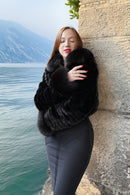 A woman with red hair and red nail polish stands by a stone wall next to a lake, wearing a luxurious black fur jacket and dark gray pants.