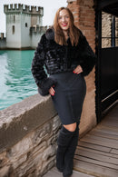 A woman posing outdoors in a black faux fur cropped jacket, a black bodycon skirt, and black over-the-knee boots, with a stone wall, brick wall, and water in the background.