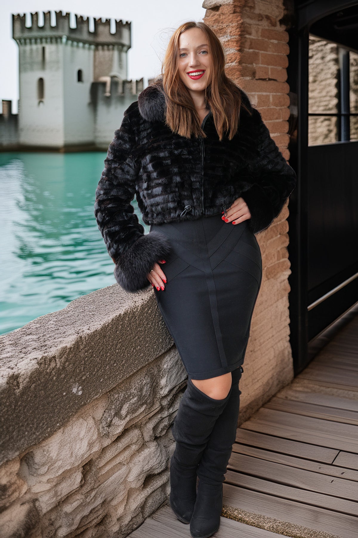 A woman posing outdoors in a black faux fur cropped jacket, a black bodycon skirt, and black over-the-knee boots, with a stone wall, brick wall, and water in the background.