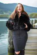 A woman stands on a wooden dock by a lake wearing a black cropped faux-fur jacket and a dark gray fitted skirt, with mountains in the background.