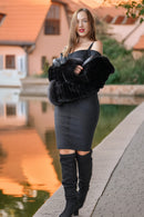 A woman stands outdoors beside a canal at sunset, wearing a black off-the-shoulder mini dress, a voluminous black faux-fur stole, and black over-the-knee boots.