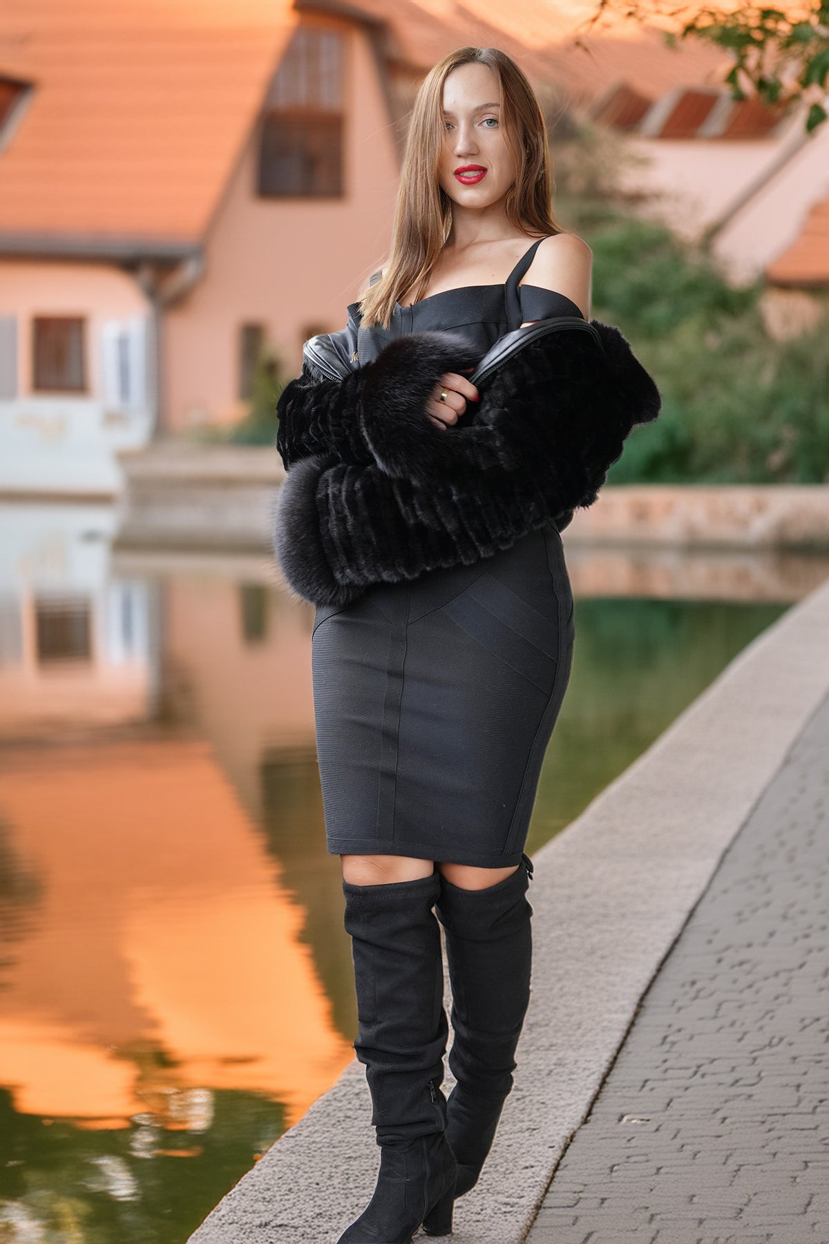 A woman stands outdoors beside a canal at sunset, wearing a black off-the-shoulder mini dress, a voluminous black faux-fur stole, and black over-the-knee boots.