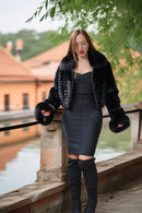 A woman stands outdoors by a stone railing next to a canal, wearing a black fur-trimmed cropped jacket over a black strapless dress and black over-the-knee boots.