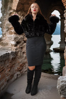 A woman stands in a stone archway by the water wearing a black faux-fur cropped jacket with fluffy cuffs, a dark gray fitted pencil skirt, and black over-the-knee boots.